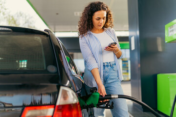 A young beautiful woman does not follow the safety rules and uses the phone while filling her car gas tank - obrazy, fototapety, plakaty