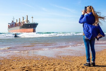 tourist films on her phone how a kite surfer surfs the waves near a large cargo ship grounded on...
