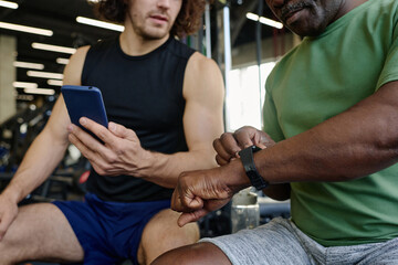 Unrecognizable caucasian athlete and unknown black man sitting at gym checking their gadgets