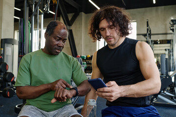 Elderly black man and young caucasian guy sitting at gym looking through their electronic devices