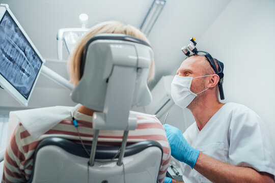Dental clinic patient visit in modern medical ward. Dentist doctor in magnifying glasses evaluating Dental X-rays scan. Patient sitting in chair. Health care and medicare industry concept image.