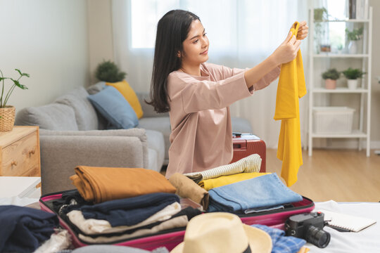 Travel concept, happy asian young woman hand preparing packing luggage bag for summer vacation trip on bed at home, pack her clothes and stuff, objects into bag suitcase for a new journey for leisure.