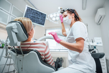 Dental clinic patient visit modern medical ward. Dentist female doctor dressed uniform in magnifying glasses pointing light. Patient sitting in chair. Health care and medicare industry concept image - obrazy, fototapety, plakaty