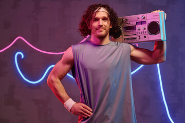 Caucasian athlete in retro sportswear posing with boombox against neon light background
