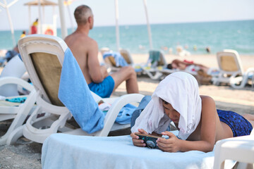 Child on a sunbed immersed in mobile gaming, beachside. Portrays modern leisure's shift from...
