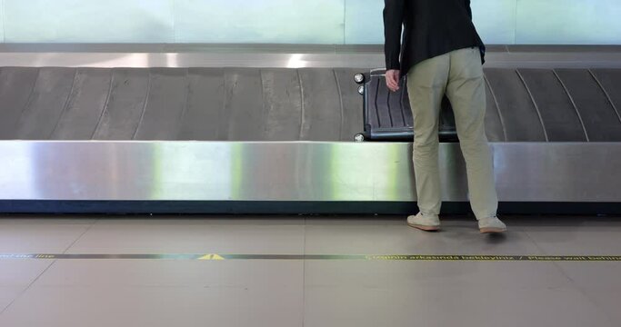 Passenger stands by baggage claim carousel in large airport, mid-shot, with focus on luggage belt. His black suitcase moves on belt, and man patiently waits for it to approach