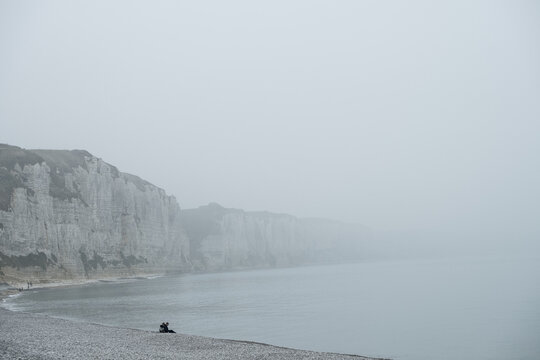 The image depicts a solitary and contemplative scene along the Fecamp coast of France, enveloped in mist. The towering white cliffs loom in the background, their grandeur softened by the foggy shroud