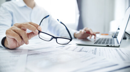 Close-up shot, businessman holding eyeglasses review many documents placed on the desk and using...