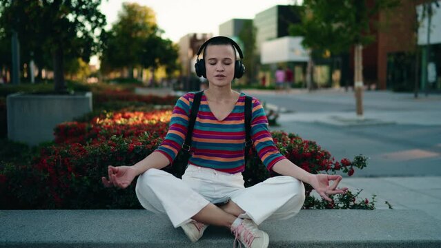 Caucasian relaxed female sits in lotus pose for meditation moment in urban setting, finding peace through online guided breathing app. Woman in headphones listening to calming music for mind control