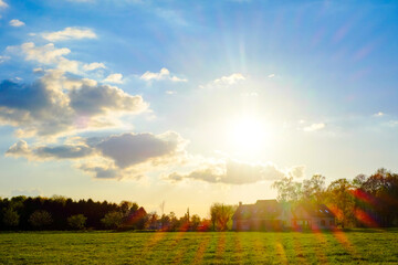 This image captures the serene beauty of a countryside sunset, with the sun's golden rays piercing through the clouds and casting a warm glow over the expansive field. The rays create a dramatic flare - obrazy, fototapety, plakaty