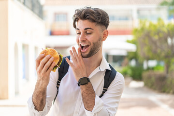 Young handsome man holding a burger at outdoors with surprise and shocked facial expression