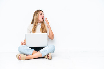 Young woman with laptop sitting on the floor isolated on white background yawning and covering wide...