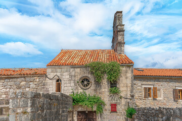 Exterior view of the Church of Santa Maria in Punta in Budva Old Town, Montenegro. Facade of an ancient stone building with a red tiled roof and a round window. Flowering caper bushes grow on the wall - obrazy, fototapety, plakaty