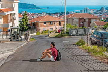 Male tourist with a backpack sits in the middle of the road on the backdrop of the cityscape of Budva and the seascape of the Adriatic Sea, Montenegro. Young man is a solo backpacker in the Balkans