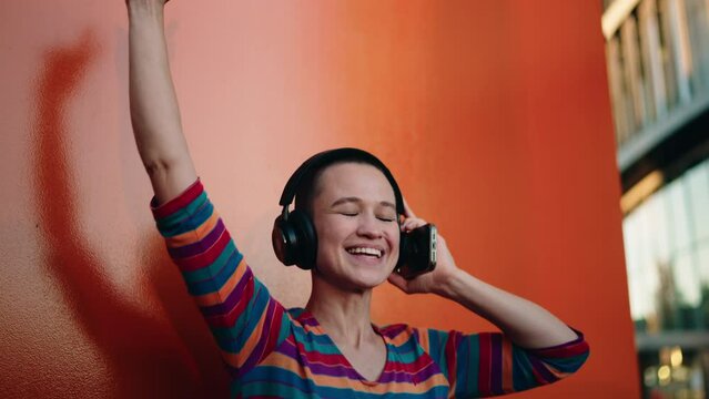 Smiling bald female in casual wear using mobile app to listen to upbeat tunes, expressing carefree spirit and happiness.Listening to music on smartphone, shadow dance on orange wall, embodying freedom