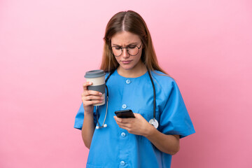 Young surgeon doctor woman isolated on pink background holding coffee to take away and a mobile