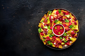 Loaded nachos. Mexican nacho chips with beef, overhead flat lay shot with guacamole sauce, cheese salsa, and chili peppers, on a black slate background, with copy space