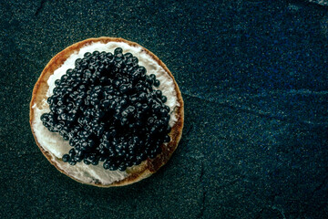 A blini with caviar and cream cheese, shot from above on a black slate background with copy space