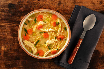 Chicken noodle soup with vegetables, a bowl of healthy broth with a spoon, overhead flat lay shot on a rustic wooden table, winter home cooking