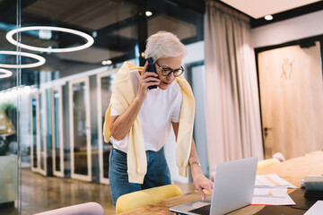 Focused senior woman standing and speaking over smartphone in modern workspace