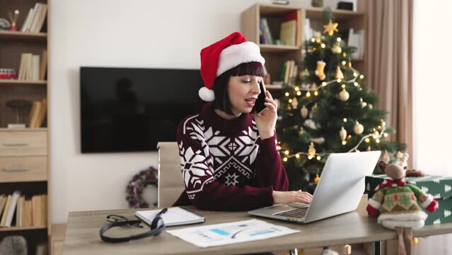 Happy woman in Santa hat talking on smartphone while using portable computer on background of Christmas tree. Efficient caucasian freelancer having business conversation with client of remote company.