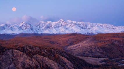 Fototapeta premium Dawn in the mountains with a full moon in the sky. Impressive panorama of dawn snow-capped peaks and a wide mountain valley in soft purple tones.