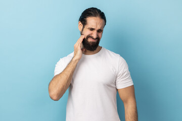 Dental problem. Portrait of man with beard wearing white T-shirt touching his cheek and grimacing...