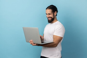 Portrait of successful man with beard entrepreneur, looking with happy smile at the camera, achieving business goals, while working on computer laptop. Indoor studio shot isolated on blue background.