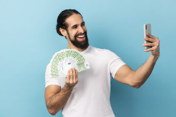 Portrait of delighted optimistic man with beard wearing white T-shirt taking selfie on cellphone with fan of euro banknotes in hands. Indoor studio shot isolated on blue background.