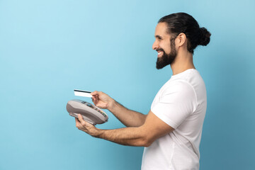 Side view of joyful man with beard wearing white T-shirt using pos payment terminal, going to make...