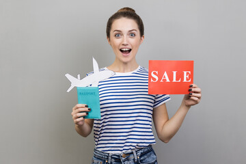 Portrait of excited woman wearing striped T-shirt holding passport with paper air plane and sale...