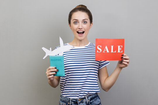 Portrait of excited woman wearing striped T-shirt holding passport with paper air plane and sale word sign, looking at camera with amazement, traveling. Indoor studio shot isolated on gray background.