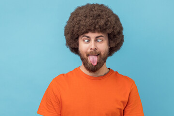 Portrait of silly man with Afro hairstyle wearing orange T-shirt making grimace and crosses eyes, playing fool, having fun alone, sticking out tongue. Indoor studio shot isolated on blue background. - obrazy, fototapety, plakaty