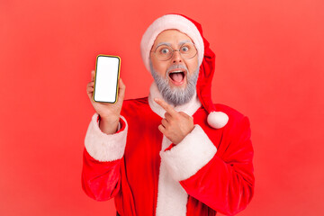 Portrait of amazed elderly man with gray beard wearing santa claus costume pointing at smart phone screen with copy space for ad, keeps mouth open. Indoor studio shot isolated on red background.