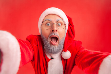 Portrait of elderly man with gray beard in santa claus costume making selfie POV or broadcasting...