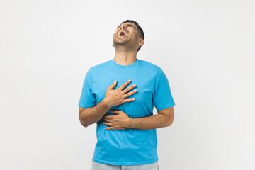 Portrait of happy optimistic crazy unshaven man wearing blue T- shirt standing listening anecdote, laughing out loud, hysterical laugh. Indoor studio shot isolated on gray background.