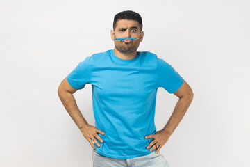 Portrait of funny childish handsome unshaven man wearing blue T- shirt standing holding pencil with his lips, pretended he has mustache. Indoor studio shot isolated on gray background. - obrazy, fototapety, plakaty