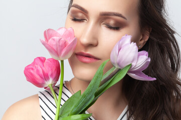 Portrait of charming cute romantic brunette woman standing with closed eyes, smelling spring tulips, enjoying fresh aromat, wearing striped dress. Indoor studio shot isolated on gray background.