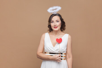 Portrait of romantic angelic woman with wavy hair and nimb over head, holding little red heart on stick, looking at camera, wearing white dress. Indoor studio shot isolated on light brown background.