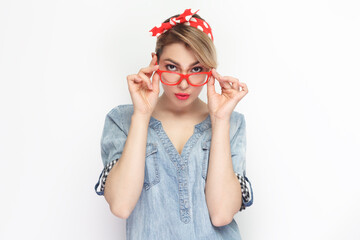 Portrait of funny beautiful attractive blonde woman wearing blue denim shirt and red headband standing looking at camera, touching frame of glasses. Indoor studio shot isolated on gray background.