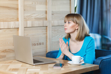 Side view of hopeful woman with blonde hair in blue shirt working on laptop, having online...