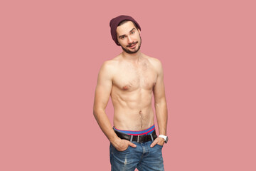 Portrait of attractive naked shirtless bearded man standing, keeps hands in pocket, looking at camera with positive flirting expression. indoor studio shot, isolated on pink background.