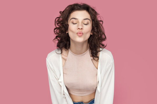 Portrait of romantic woman with curly hairstyle wearing casual style outfit keeps eyes closed and sending air kiss, flirting with boyfriend. Indoor studio shot isolated on pink background.