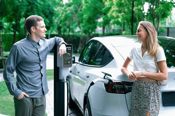 Young couple travel with EV electric car charging in green sustainable city outdoor garden in...