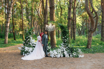 Beautiful couple, the groom in a green suit and the bride in an unusual pink wedding dress, posing at a luxurious ceremony with beautiful decor and an arch with a chandelier and candles in the forest 