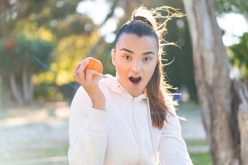 Young pretty brunette woman holding an orange at outdoors with surprise and shocked facial expression