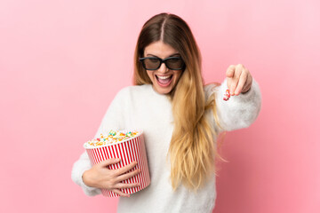 Obraz premium Young blonde woman isolated on pink background with 3d glasses and holding a big bucket of popcorns while pointing front