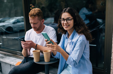 Portrait of happy Caucasian female blogger in optical spectacles for vision correction smiling at camera while millennial boyfriend browsing website on modern smartphone device, technology users