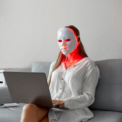 A beautiful girl with an LED mask on her head works at home on a laptop. Home skin care concept.