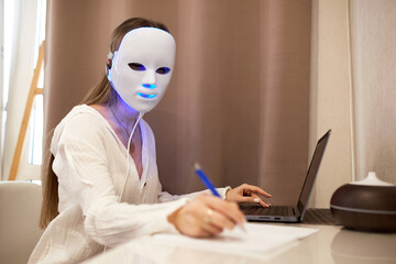 A beautiful girl with an LED mask on her head works at a laptop. Home skin care concept.